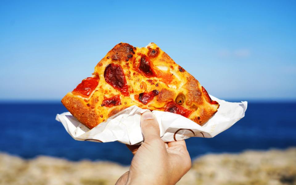 Focaccia con pomodori e olive, tenuta in mano, sfondo mare e cielo blu.