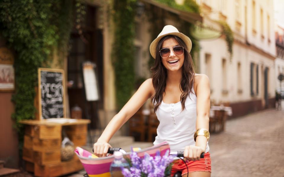 Donna sorridente in bicicletta, indossa cappello e occhiali da sole su strada cittadina.