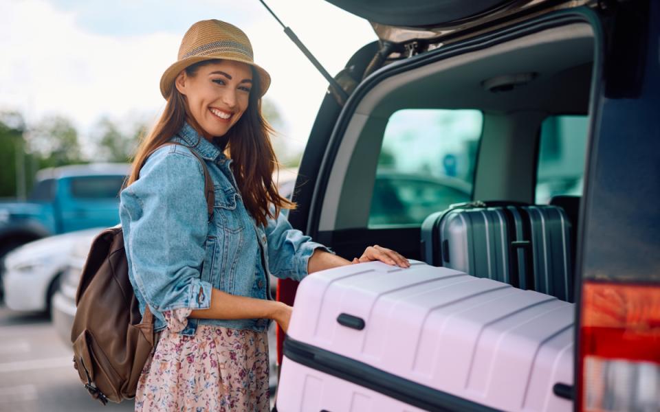 Donna felice carica valigia in auto, indossa cappello e giacca di jeans.